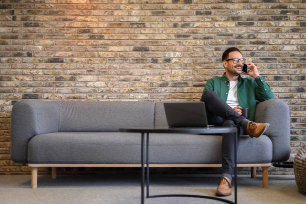 Happy male programmer with coffee cup discussing ideas over phone call while sitting on sofa in office