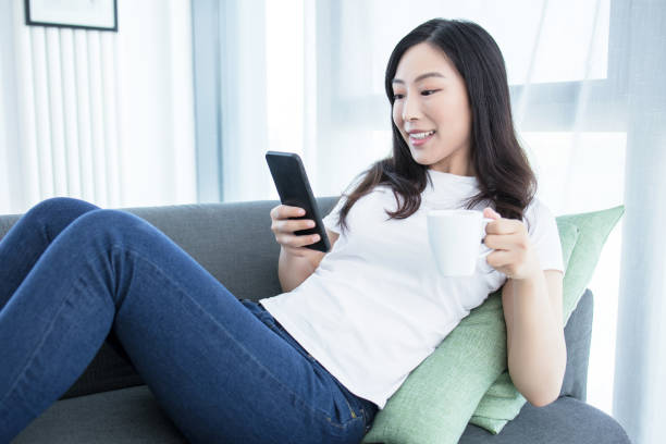 istock Smiling woman sitting on sofa holding coffee using phone 2171996432