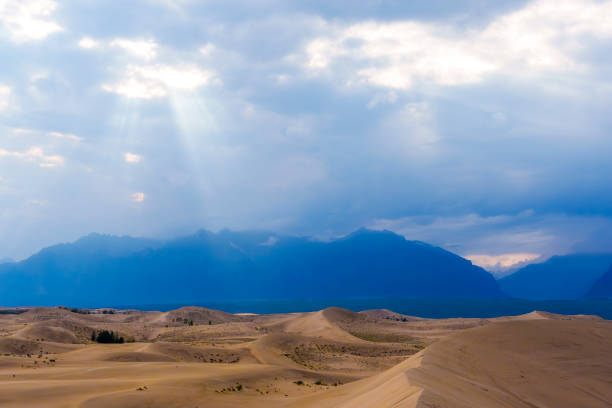Expansive desert dunes stretch under a dramatic sky with sunbeams breaking through the clouds, creating a serene and awe-inspiring landscape.