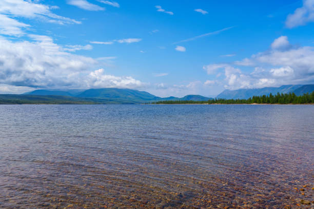 A clear lake with a gently sloping rocky shore, surrounded by lush green forests and distant mountains under a partly cloudy sky, creating a serene and picturesque landscape.
