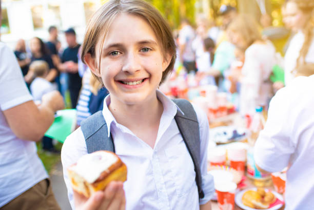 A boy in a school uniform and with a backpack in his hands holds a bun while standing in front of a festive table at a tea party at school