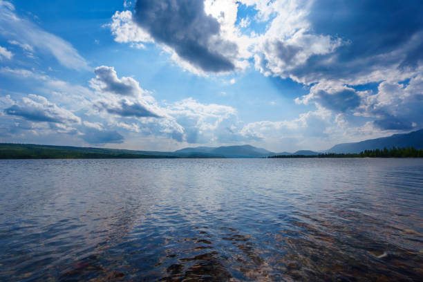 A tranquil lake with smooth waters reflects the soft light of a misty morning, framed by distant mountains and a tree-lined shore, creating a serene and peaceful landscape.