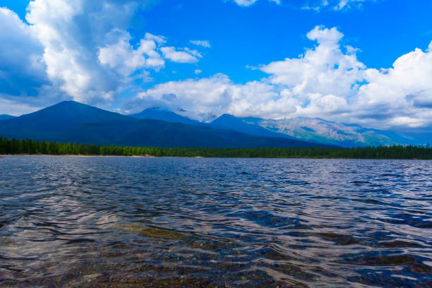 A clear lake with a gently sloping rocky shore, surrounded by lush green forests and distant mountains under a partly cloudy sky, creating a serene and picturesque landscape.