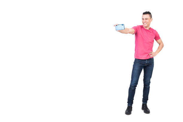 Full length of self assured young happy male with trendy haircut in casual outfit standing against white background with hand on waist and taking selfie on smartphone