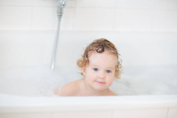 Little baby in bath tub playing with soap foam. Toddler girl brushing teeth and washing hair with shampoo. Shower wash for infant. Mother bathing baby.