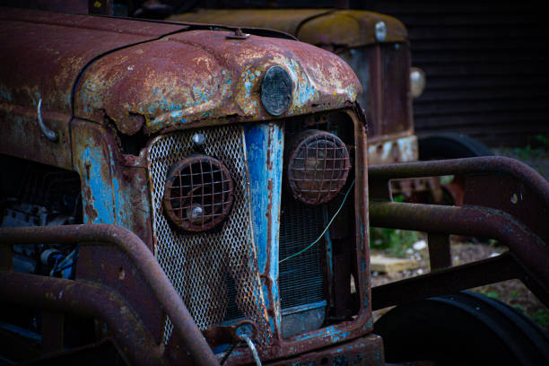 Old, rusted tractor parked in an overgrown farmyard with dilapidated sheds in the background under a blue sky.