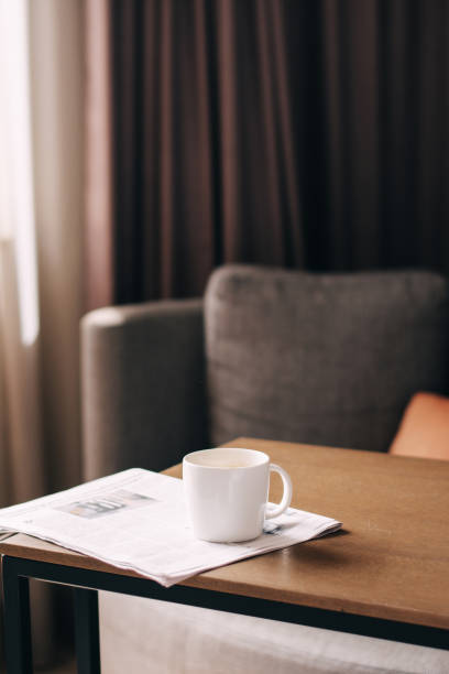 A cup of coffee is on the table. There is a newspaper nearby. The morning sun is shining through the window.