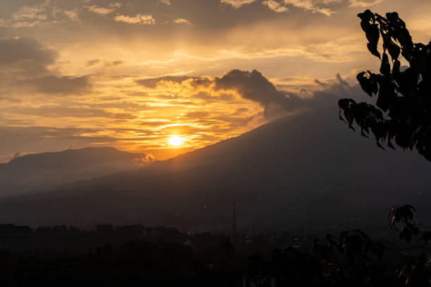 This image captures a beautiful sunset over a mountain range. The sky is a vibrant orange and yellow, with cloudy sky. The sun is setting behind the mountains, casting long shadows over the landscape.