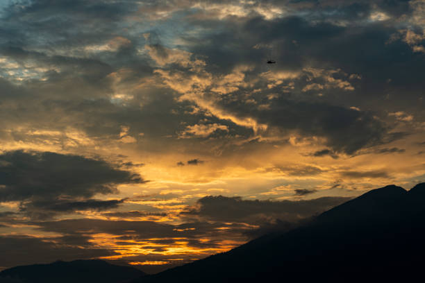 This image captures a beautiful sunset over a mountain range. The sky is a vibrant orange and yellow, with cloudy sky. The sun is setting behind the mountains, casting long shadows over the landscape.