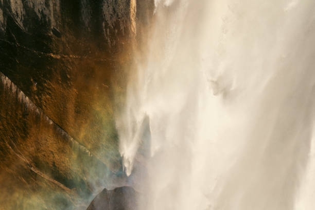 This stunning close-up captures the mesmerizing textures of Yosemite Falls, where the cascading water forms what appears to be a face-like structure within the falls. The thick white veil of water rushes down with immense force, illuminated by sunlight that creates a striking rainbow on the left side of the image. The interplay of light, water, and nature gives the falls a magical, almost mystical appearance. The rainbow enhances the dreamlike quality of this natural wonder, creating a unique moment of beauty and serenity in Yosemite National Park.