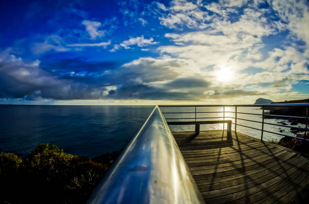 A bench is on a pier overlooking the ocean. The sky is blue with some clouds. The sun is setting, casting a warm glow on the water