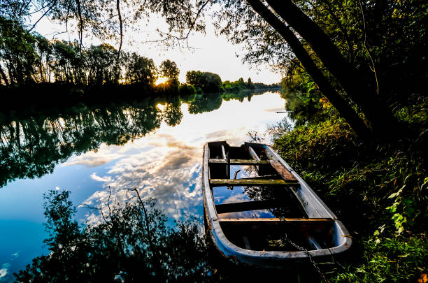 A boat is sitting in the water next to a river. The sky is cloudy and the sun is setting