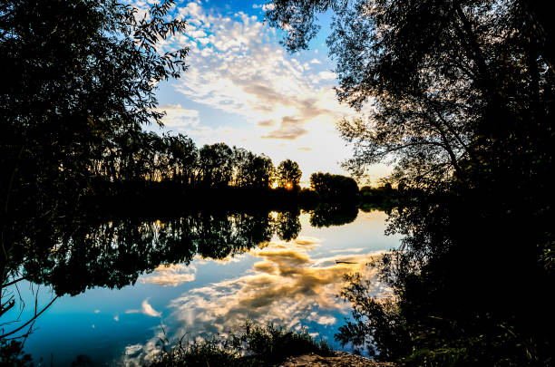 A serene lake with a beautiful sunset in the background. The water is calm and the sky is filled with clouds. The trees surrounding the lake are tall and provide a sense of tranquility