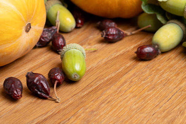 A collection of acorns and dried berries scattered on a wooden surface alongside a yellow squash. The arrangement highlights the natural elements of autumn harvest and seasonal produce.