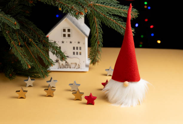 A festive Christmas scene featuring a small gnome with a red hat, a white house ornament, and wooden stars scattered around. Evergreen branches and soft colorful lights in the background enhance the holiday atmosphere.