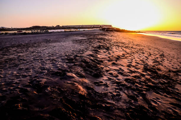 A beach with a sunset in the background. The beach is covered in sand and there are a few chairs scattered around