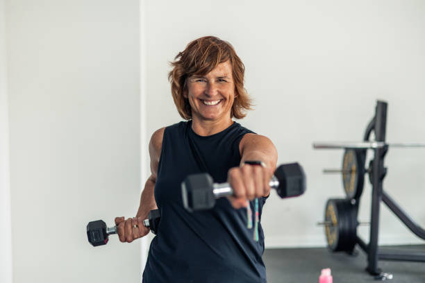 Elderly woman smiling at camera while doing arm exercises with dumbbells during gym workout