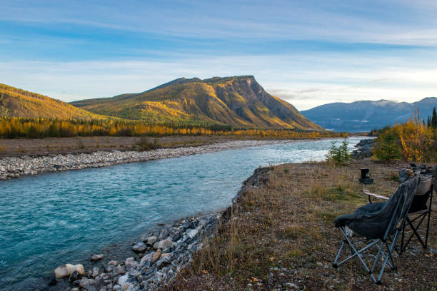 Golden and yellow autumn colours are visible on mountains lit by the rising sun across a flowing river in northern British Columbia, Canada. Camping chairs and coffee maker are visible in the bottom right corner at this overlanding and boondocking site on a riverbank in a remote location.