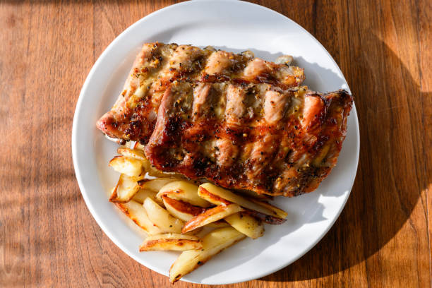 Marinated pork ribs and young boiled potatoes in a wide ceramic plate on a wooden table, next to them are a table fork and knife. Close-up.
