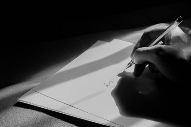 The figure of a hand writing on a white paper. The writing activity is illuminated by light from outside the window.