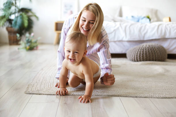 Cute cheerful European infant in diaper having joyful facial expression, laughing while crawling on floor from his smiling mother who is chasing him. Blonde young woman playing with son indoors