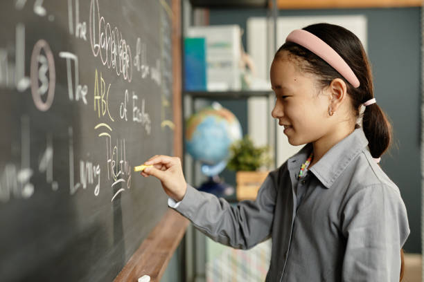 istock Smiling Young Girl in Headband Writing on Blackboard during Lesson in Classroom 2172310329