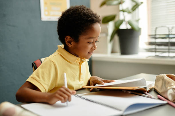 istock Smiling Black Boy Sitting at Desk Reading Textbook while Studying in Class at School 2172310342