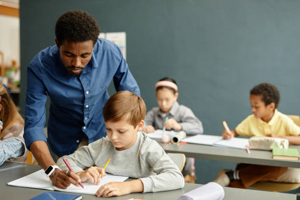 istock Male Teacher Helping Young Boy with Spelling while Writing in Copybook at School 2172310343
