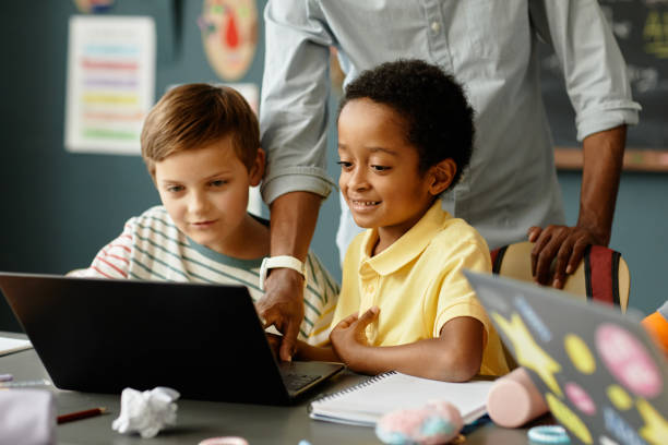 istock Smiling Black Boy Using Laptop while Teacher Helping during Lesson at School 2172310395