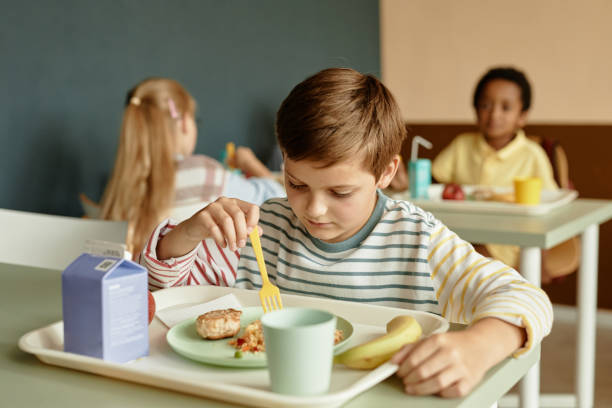 istock Boy Sitting at Table Looking at Plate Eating Lunch in School Cafeteria 2172310461