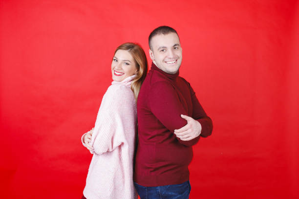 Young heterosexual couple in turtleneck sweaters standing back to back and smiling at camera with joy, red colored background, studio shot