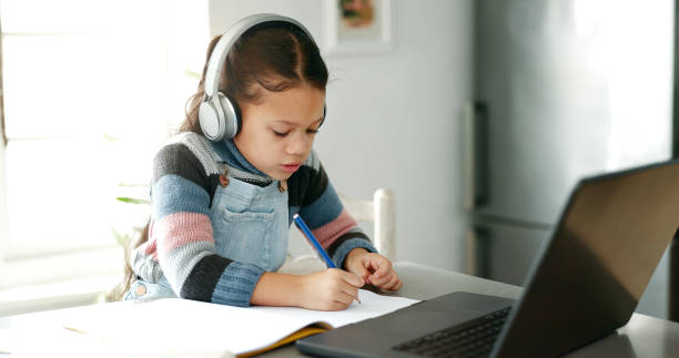 istock Home, girl and homework with laptop for education, elearning and connection for online class. Child, studying and cognitive development with writing for learner, knowledge and headphones in kitchen 2172335758