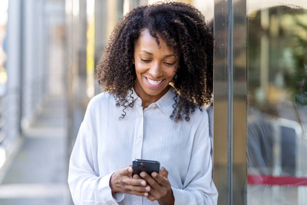 istock Curly haired businesswoman smiling and using smartphone while leaning against office building 2172342889