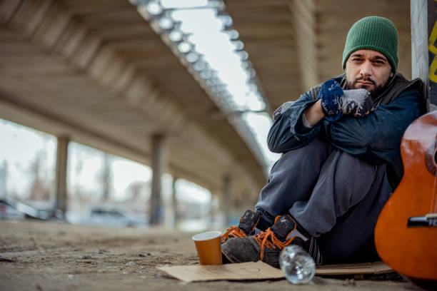 Mid Adult Homeless Man Looking at Camera While Sitting Under the Bridge. Homelessness Social Issues Concept.