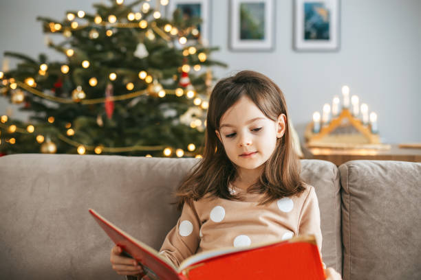 istock A little girl reading a big red book sitting on a sofa, while a Christmas tree stands behind her 2172351634