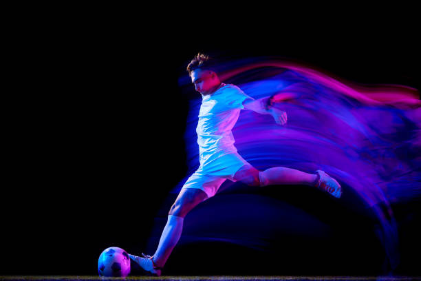 Focused football player, dressed in white under neon lighting, performs swift mid-air kick, mastering agility and technique in his practice. Motion blur.