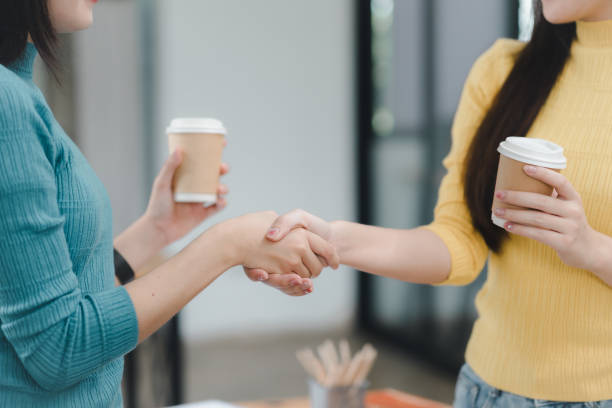 istock Two women shaking hands while holding coffee cups, showcasing friendly business interaction. 2172363953