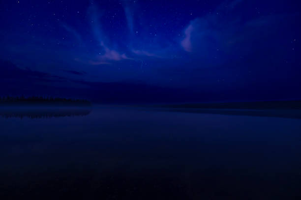 A dark, starry night sky with scattered clouds reflected on the surface of a tranquil lake.