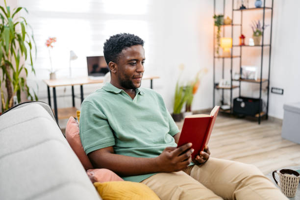 istock Young Man Reading A Book While Sitting On The Sofa At Home 2172367546