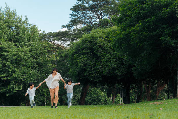 istock A mother and her two young children walk together outdoors under large trees, holding hands and smiling. A warm family moment in nature, captured in a sunny park setting. 2172378455
