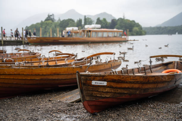 A wooden boat rests at a small pier on Derwentwater, as a flock of ducks glides across the gently rippled lake, with a backdrop of distant fells under a cloudy summer sky