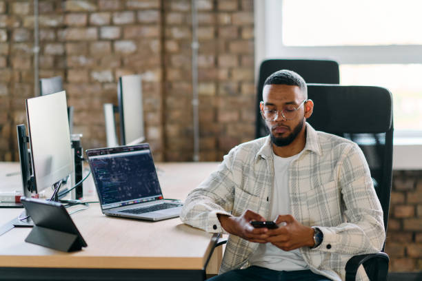 African American entrepreneur takes a break in a modern office, using a smartphone to browse social media, capturing a moment of digital connectivity and relaxation amidst his business endeavors.