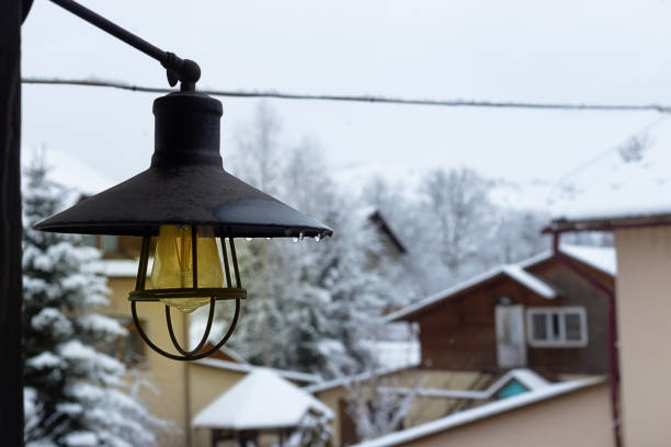 A street black lantern covered in snow, with a white paper cup with a black lid on top. In the background, a wooden building
