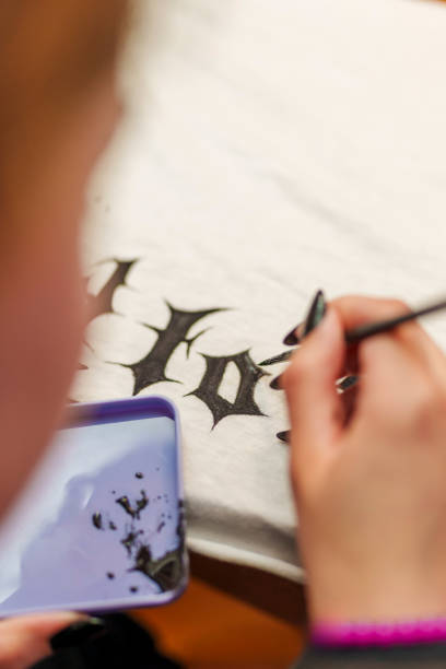 The artist uses a fine brush to paint precise gothic letters on fabric, capturing the dedication to detail in the creative process.