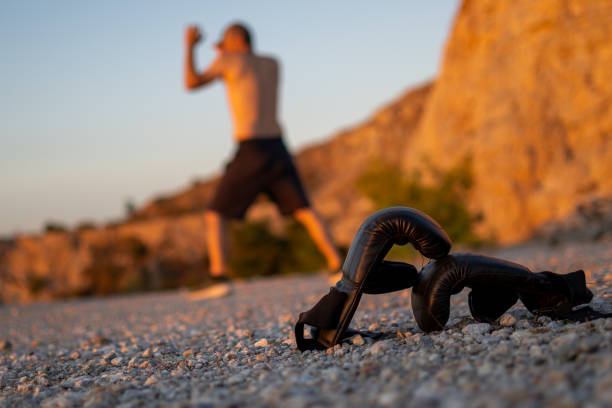 Photo shows a person with no t-shirt training shadow boxing outdoors. Selective focus at the boxing gloves on the ground.