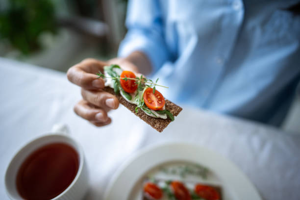 istock Hands hold rye crispbread sandwich with cream cheese, tomato, arugula, basil with tea at table 2172451977