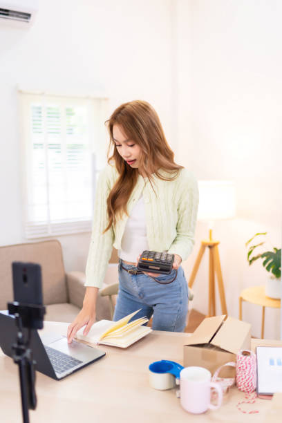 istock A young woman is standing at her desk, using her phone while organizing her workspace for her online clothing business. She is filming content, with packaging supplies and clothing around her 2172516619