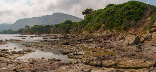 A wide-angle panoramic view of a rugged coastal landscape featuring rocky shores, lush green cliffs, and distant hills under a partly cloudy sky. Ideal for nature and travel photography