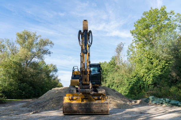 A large excavator is actively moving dirt on a construction site, surrounded by a vibrant landscape of trees and blue sky, showcasing heavy machinery in action.