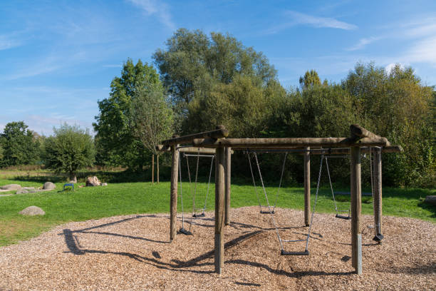 A wooden swing set sits on a bed of pebbles in a tranquil park, surrounded by lush greenery and trees under a clear blue sky. Perfect for a sunny afternoon outing.